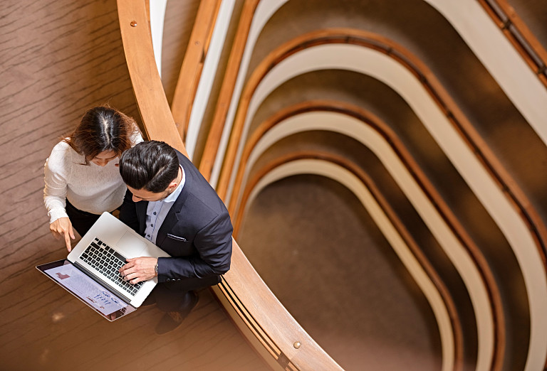 Two business people (man and woman) standing by the stairs in an office building, looking at a laptop