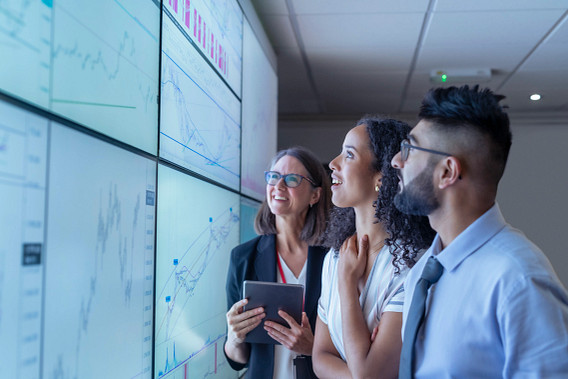 Three business professionals stand together looking at large digital screens displaying charts and graphs. One woman holds a tablet while the group smiles, appearing to analyze data or discuss results in a modern office. entrants