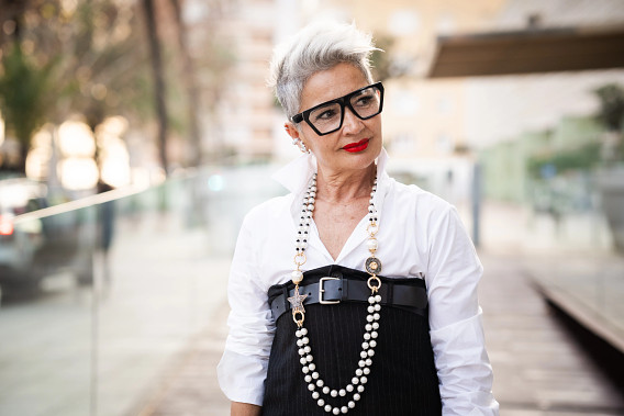 A stylish older woman with short gray hair and bold red lipstick stands outdoors, wearing a white collared shirt, a black belted dress, large black glasses, and a long pearl necklace. The background features a blurred urban setting with glass railings and greenery.