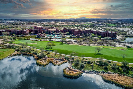 Aerial view of the Sheraton Grand at Wild Horse Pass in Phoenix, Arizona at twilight time. Behind the hotel building there is mostly desert land, with patchy greenery and mountains in the distance. In front of the building in the hotel yard, plus a green field and a pond