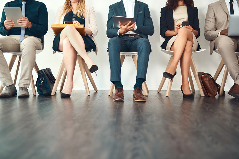 A row of five professionally dressed people sit on chairs in a waiting area, holding folders, tablets, and phones, with their legs and lower bodies visible against a dark wooden floor.