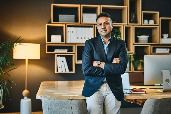 A man wearing a blue suit jacket leaning on a wooden work desk, with his hands crossed across his chest. There are square wooden shelves in the background, along with a lamp and a plant